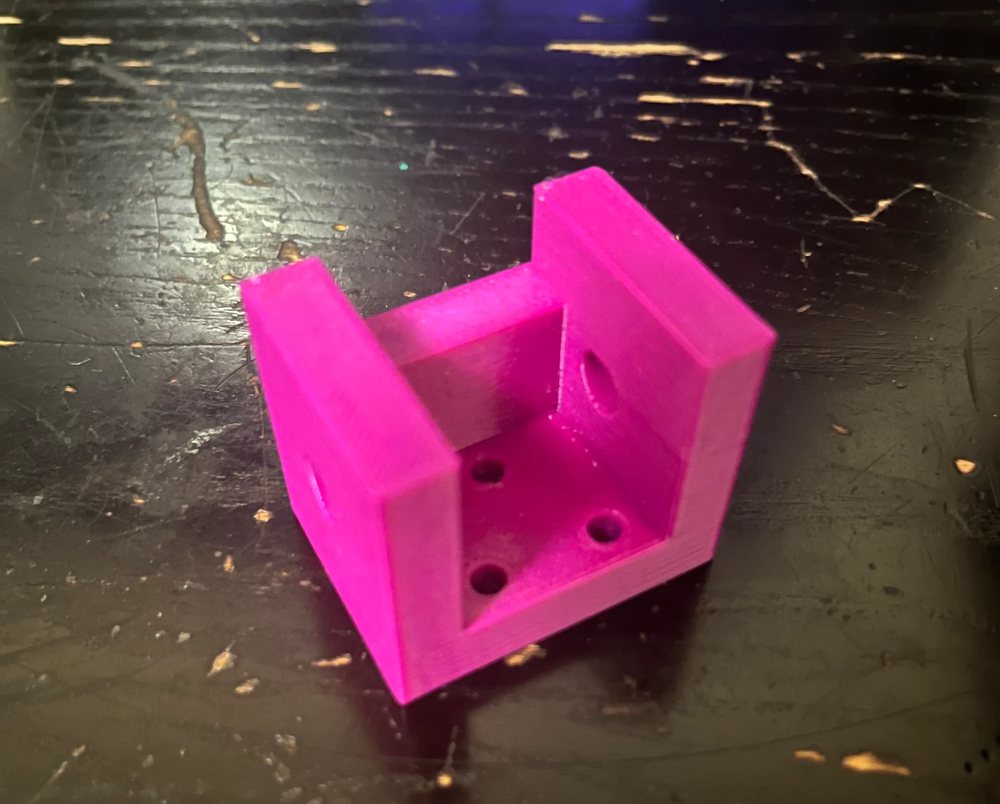 A magenta 3D-printed U-shaped bracket with four mounting holes in its base and a hole in each side wall.