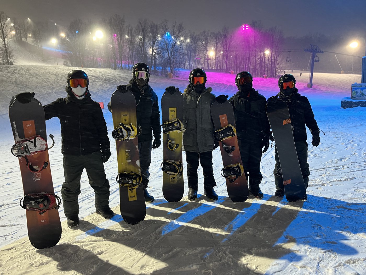 Five friends in helmets and goggles holding snowboards on a lit ski hill at night.